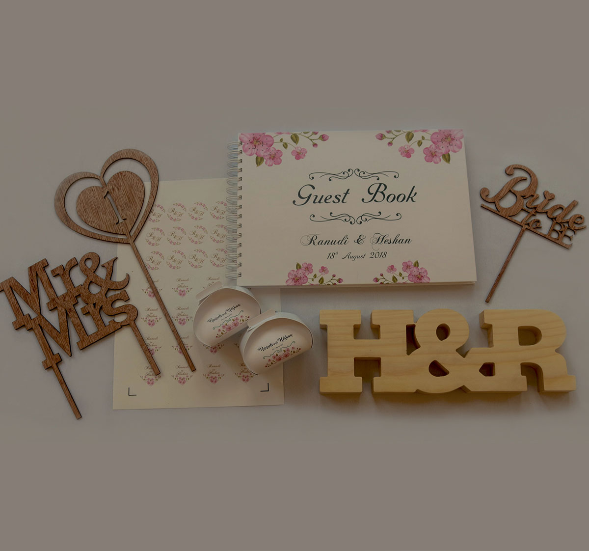 This image contains a cake topper that says Mr. & Mrs, a cake topper that says Bride to Be, a spiralbound wedding guest book, 2 cake boxes, a  wooden table number sign, a table word block that says H&R, and wedding invitation envelope sticker sheet all printed with a floral design.